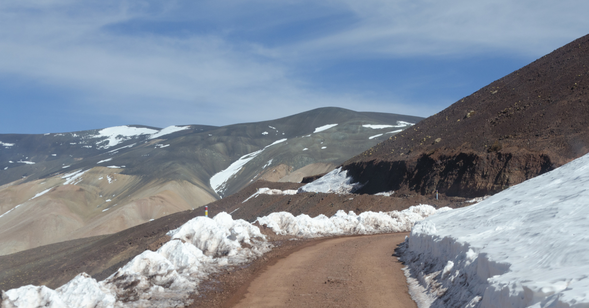 A dirt road leads through a snowy landscape, with a mountain partially blanketed in snow rising in the distance.